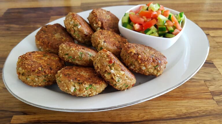 A white plate holding several golden-brown falafel patties arranged in a semicircle, with a small bowl of chopped salad consisting of tomatoes, cucumbers, and green peppers on the side. The plate is set on a wooden tabletop.