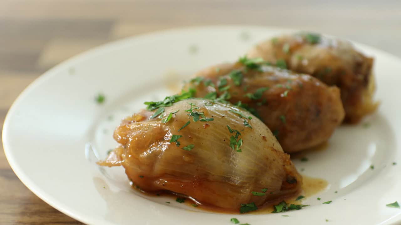 A white plate holds three stuffed onion rolls topped with a light sauce and garnished with chopped parsley. The rolls are neatly arranged in a row, showcasing their slightly caramelized and glazed appearance. The background shows a blurred wooden surface.