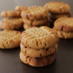 A close-up shot of stacks of freshly baked peanut butter cookies. The cookies have a golden brown color with a criss-cross pattern on top, all arranged on a dark wooden surface.