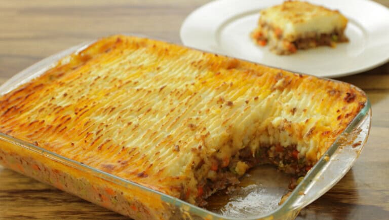 A glass baking dish filled with baked shepherd's pie, showing a layer of golden-brown mashed potatoes on top with visible ground meat and vegetables underneath. A piece has been cut out and sits on a white plate in the background.