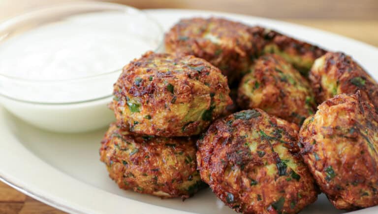 A plate of golden-brown fritters garnished with herbs, served alongside a small bowl of white dipping sauce. The fritters appear crispy and well-seasoned. The background is a blurred wooden surface.