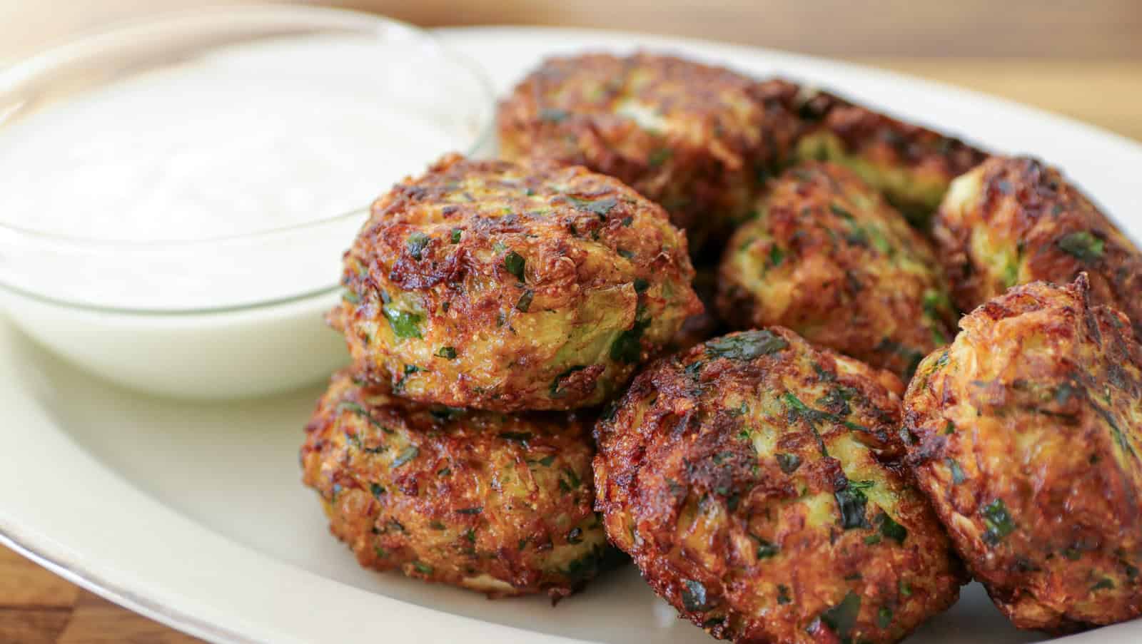 A plate of golden-brown fritters garnished with herbs, served alongside a small bowl of white dipping sauce. The fritters appear crispy and well-seasoned. The background is a blurred wooden surface.