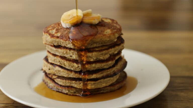 A stack of six pancakes topped with sliced bananas and drizzled with syrup on a white plate. The syrup is flowing down the sides of the pancakes. The background is a wooden table.