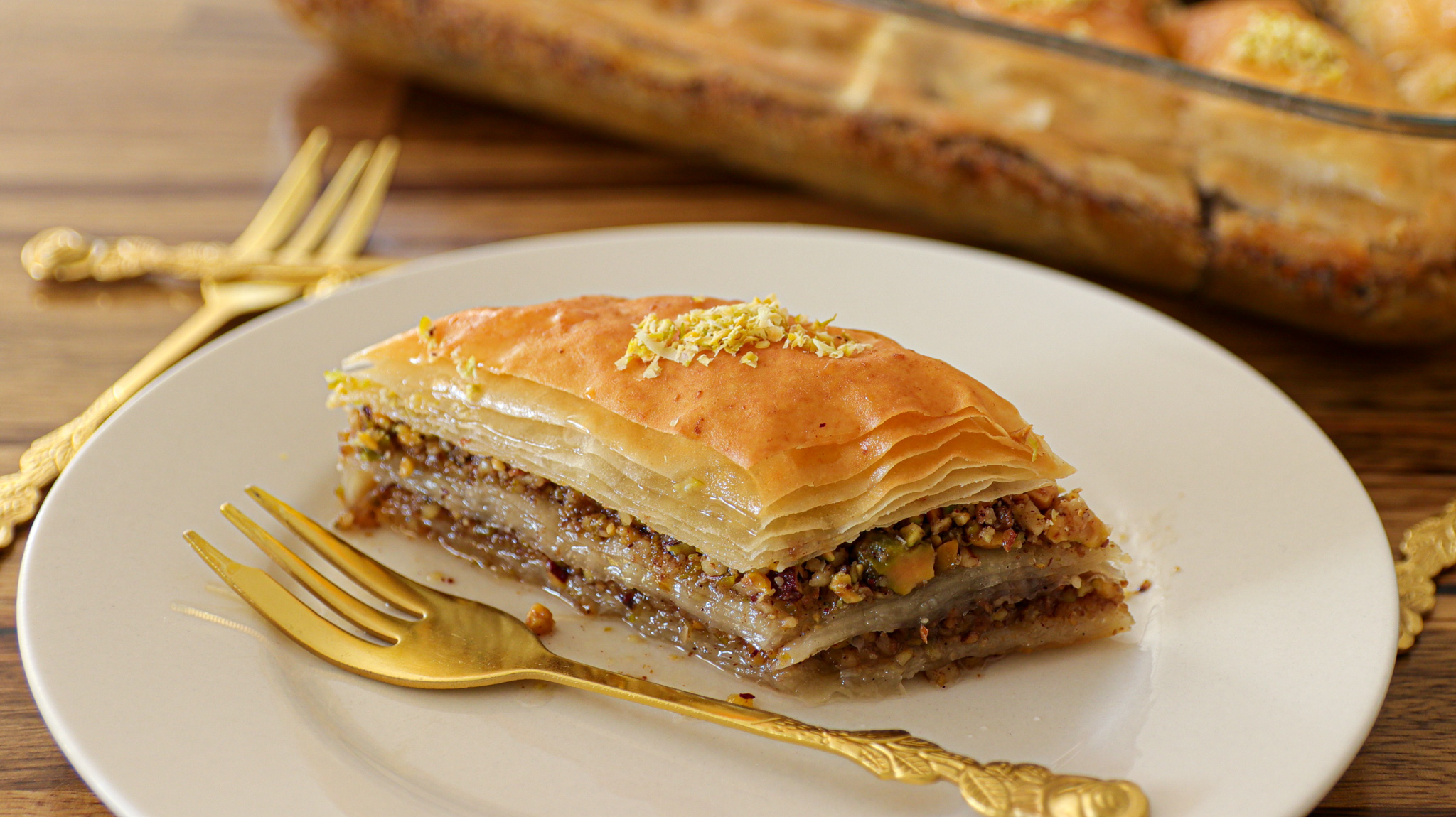 A glass baking dish filled with sliced pieces of golden-brown baklava sits on a wooden table. A triangular piece of baklava, garnished with crushed nuts, is served on a white plate in the foreground, accompanied by a small gold fork.