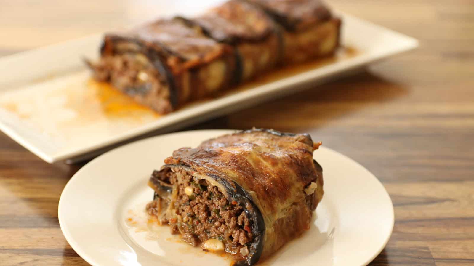 A knife slicing through a rectangular loaf of meatloaf on a white rectangular plate. The meatloaf has a caramelized crust and is covered in a rich brown sauce. The background features a wooden surface.