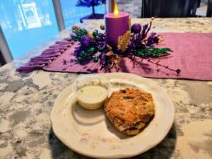 A crab cake with a small cup of dipping sauce on a floral plate, set on a marble table with a purple candle and festive holiday centerpiece in the background.