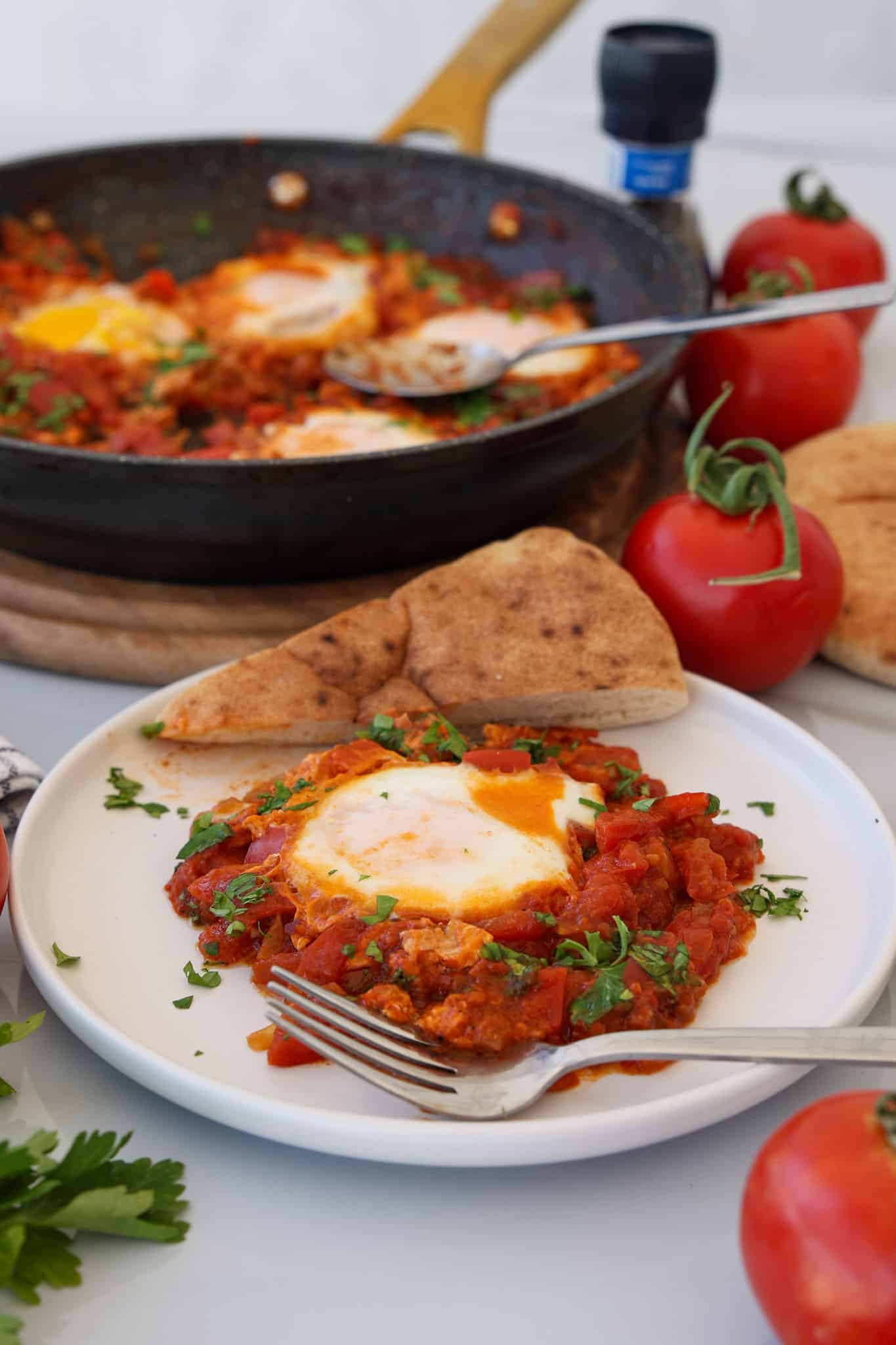 A plate of shakshuka with a poached egg in spicy tomato sauce, garnished with herbs, served with pita bread. In the background, a pan with more shakshuka and whole tomatoes are visible.