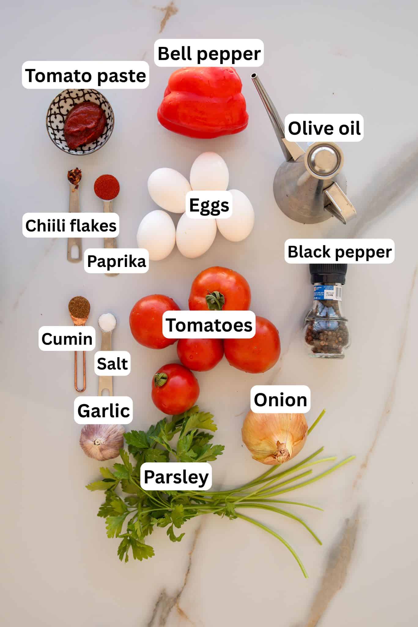 A flat lay of shakshuka ingredients on a white surface: tomato paste, bell pepper, olive oil, eggs, chili flakes, paprika, black pepper, cumin, salt, tomatoes, garlic, parsley, and onion, all labeled.