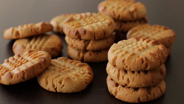 A close-up shot of stacks of freshly baked peanut butter cookies. The cookies have a golden brown color with a criss-cross pattern on top, all arranged on a dark wooden surface.