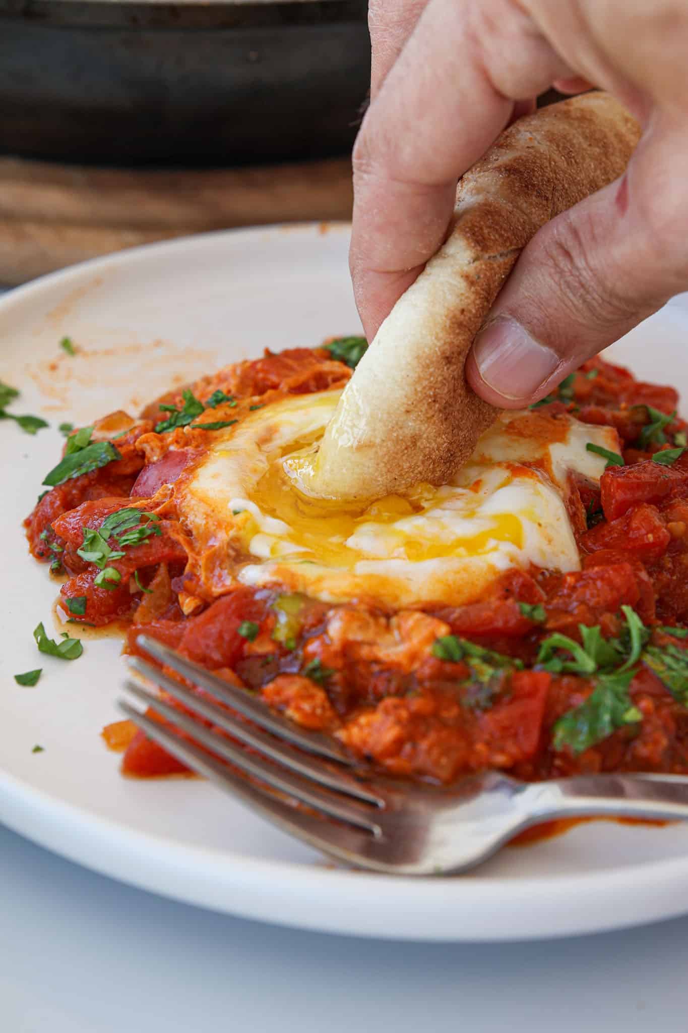A hand dips bread into a runny egg yolk on a plate of shakshuka, a tomato and pepper stew garnished with chopped herbs. A fork rests on the white plate.