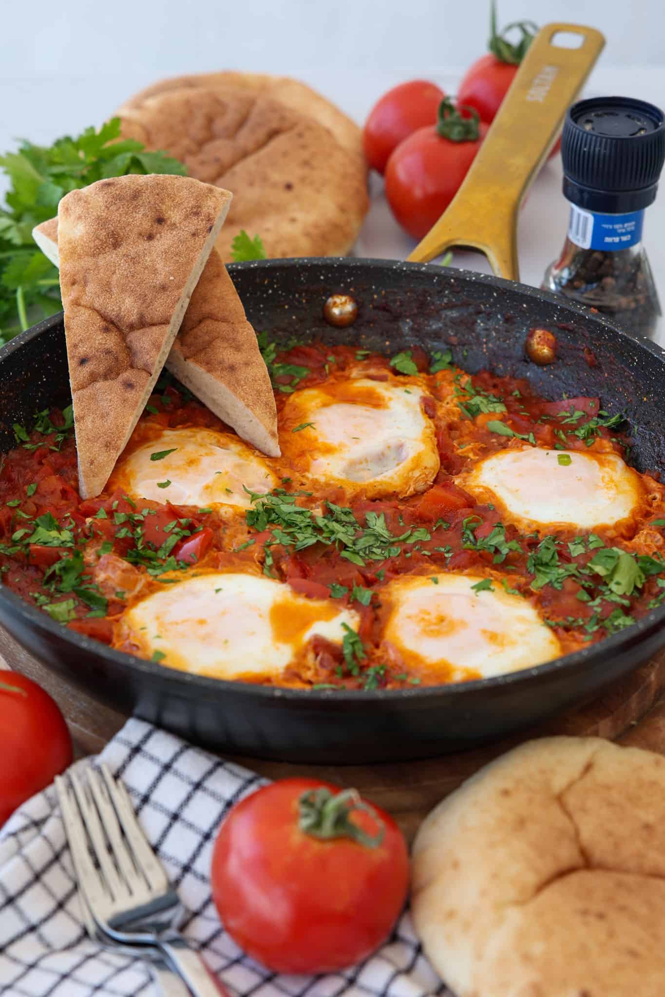 A skillet of shakshuka with poached eggs in a tomato sauce, topped with chopped herbs, surrounded by pita bread, fresh tomatoes, and a pepper grinder. Two pita wedges are propped on the pan's edge.