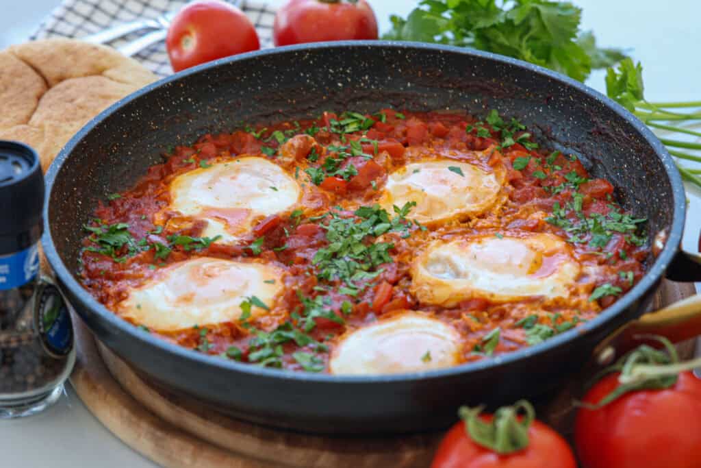 A skillet of shakshuka with poached eggs in a tomato and pepper sauce, garnished with fresh herbs, sits on a wooden board with tomatoes, herbs, bread, and a pepper grinder nearby.