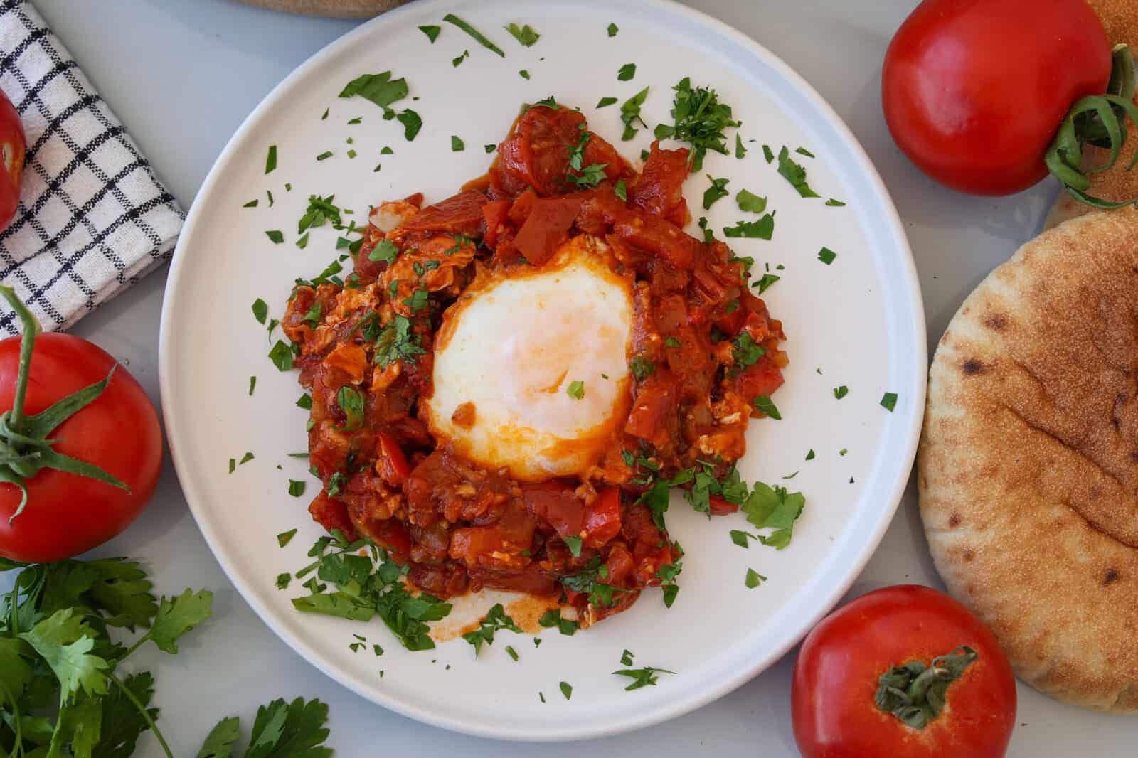A plate of shakshuka with a poached egg in the center, surrounded by a tomato, pepper, and herb sauce, garnished with fresh parsley. Fresh tomatoes, pita bread, and herbs are placed nearby.