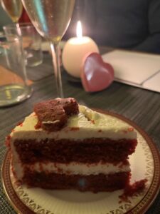 A slice of red velvet cake with white frosting and a heart-shaped brownie on top sits on a decorative plate. A lit candle, heart-shaped object, glasses, and a champagne flute are on the table in the background.