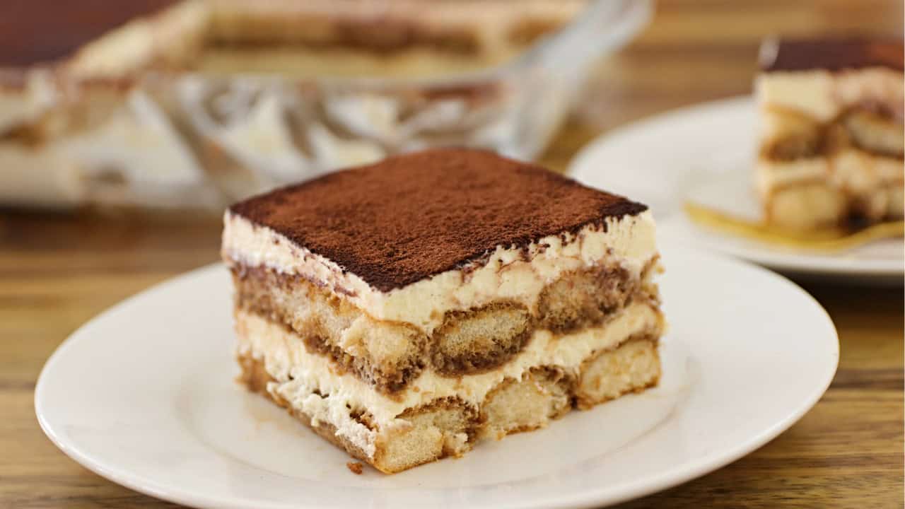 A slice of tiramisu is served on a white plate. The dessert features visible layers of coffee-soaked ladyfingers and mascarpone cream, topped with a dusting of cocoa powder. Another slice of tiramisu and the dessert tray are blurred in the background.