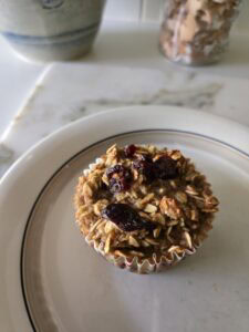 A single baked oatmeal muffin topped with dried cranberries sits on a white plate with a thin brown rim. The background includes a marble countertop and a blurred jar.