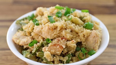 A bowl of cauliflower rice mixed with pieces of chicken and green beans, garnished with chopped green onions, is placed on a wooden surface. The dish has a textured, crumbly appearance with visible chunks of chicken and vegetables.