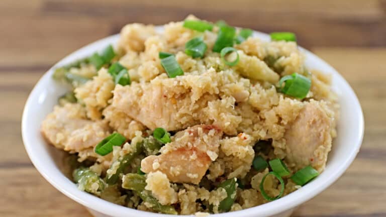 A bowl of cauliflower rice mixed with pieces of chicken and green beans, garnished with chopped green onions, is placed on a wooden surface. The dish has a textured, crumbly appearance with visible chunks of chicken and vegetables.