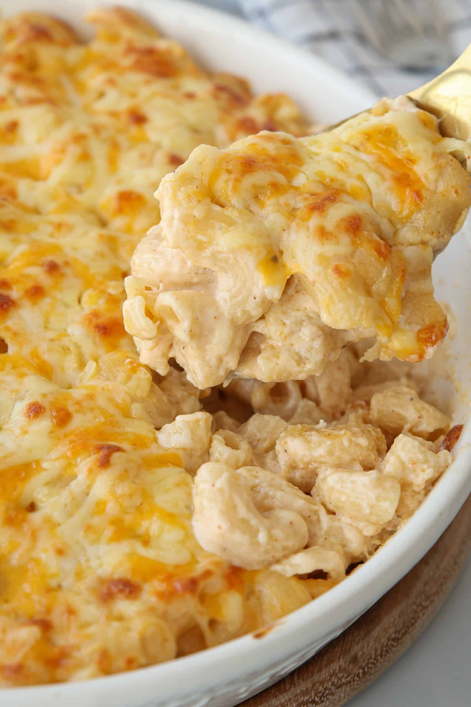 A close-up of a cheesy baked pasta dish, with a spoon lifting a creamy, golden-brown serving of pasta covered in melted cheese from a white casserole dish.
