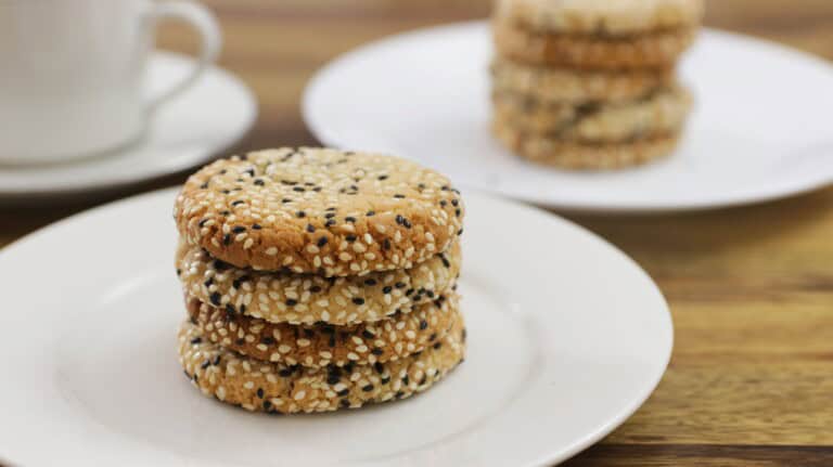 A stack of four sesame seed cookies rests on a white plate. In the background, another plate with more stacked cookies and a white cup are slightly blurred on a wooden surface.