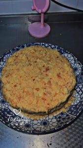 A homemade layered cake topped with crumbled pieces is presented on a decorative blue and white plate. In the background, there is a small pink mirror on a metallic countertop. The text “Tuesday” appears on the image.