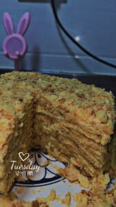 A layered cake with a slice removed sits on a plate. In the background, there’s a pink bunny-ear decoration. The text overlay reads “Tuesday 12:44 AM” with a heart graphic above the word Tuesday.
