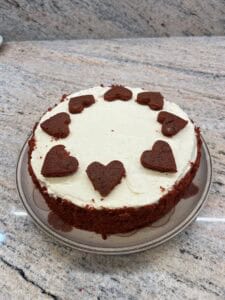 A round cake with white frosting, decorated with heart-shaped red velvet pieces around the edge, sits on a glass plate on a light granite countertop.