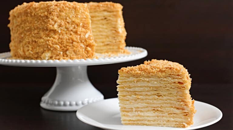 A multi-layered honey cake is displayed on a white pedestal cake stand against a dark background. A separate slice of the cake sits on a white plate in the foreground, showing its numerous thin layers and golden, crumb-coated exterior.