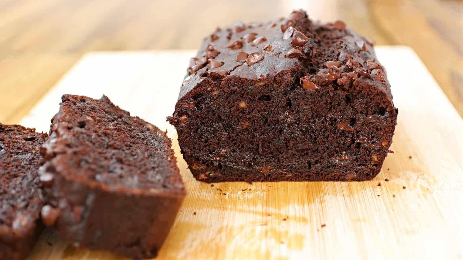 A loaf of chocolate chip banana bread lies on a wooden surface. One slice is cut and positioned next to the loaf, showing the moist, rich chocolate interior dotted with chocolate chips. The background is softly blurred.