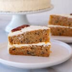 A close-up of a slice of carrot cake on a white plate. The cake has two layers of moist, brown cake with visible nuts and is filled and topped with white cream cheese frosting. Another slice of cake is blurred in the background.