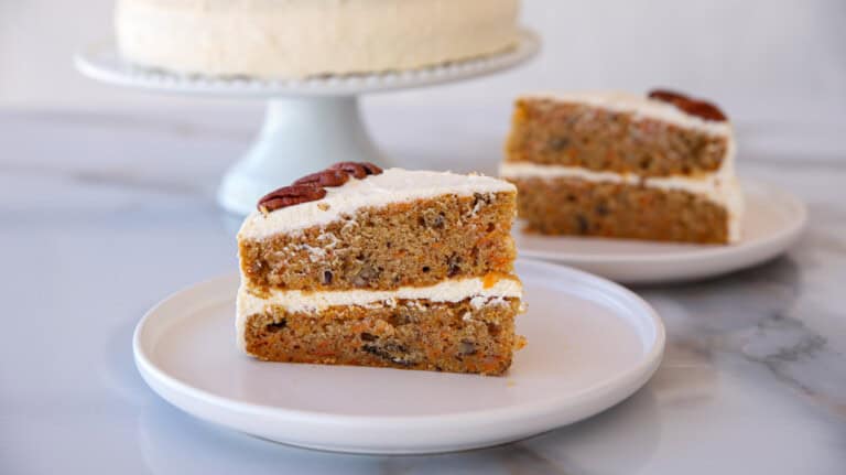 A close-up of a slice of carrot cake on a white plate. The cake has two layers of moist, brown cake with visible nuts and is filled and topped with white cream cheese frosting. Another slice of cake is blurred in the background.