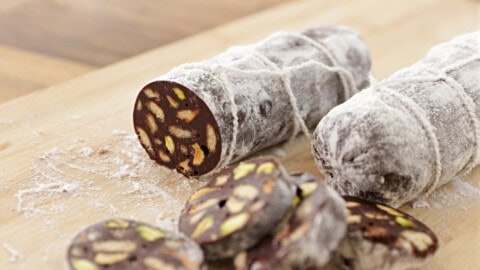 Two chocolate salami logs covered in powdered sugar are displayed on a wooden cutting board. One log is shown sliced, revealing the interior which contains visible chunks of nuts and biscuits. Several round slices are placed in front of the log.