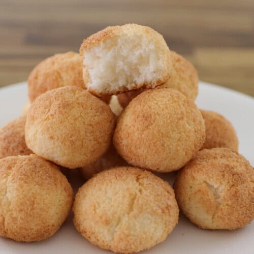 A white plate with a pyramid of golden-brown coconut macaroons. The macaroon on the top has a bite taken out, revealing a fluffy interior. The background is a wooden table.