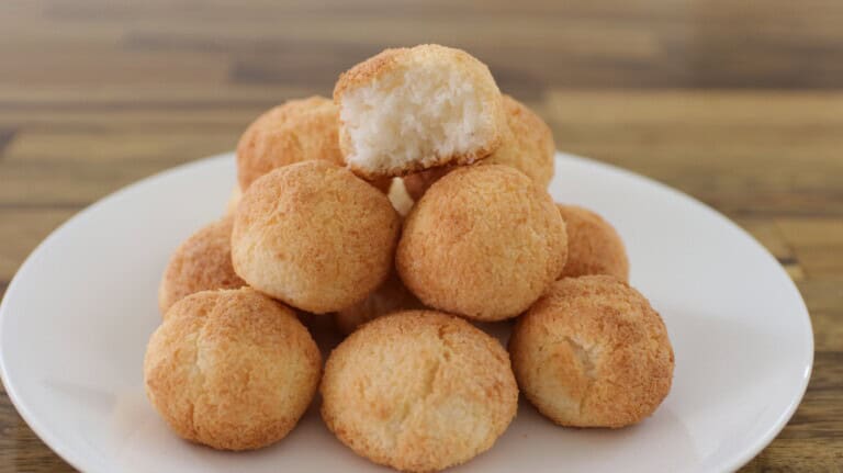 A white plate with a pyramid of golden-brown coconut macaroons. The macaroon on the top has a bite taken out, revealing a fluffy interior. The background is a wooden table.