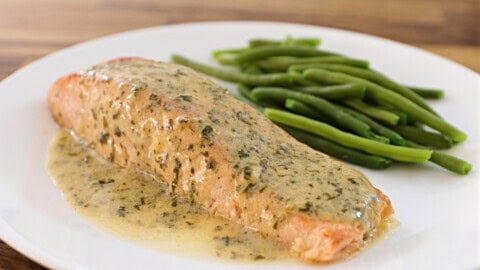 A white plate featuring a cooked salmon fillet topped with a creamy herb sauce, accompanied by a serving of steamed green beans. The dish is presented on a wooden surface.