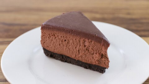 A slice of rich chocolate cheesecake with a dark chocolate crust sits on a white plate. A golden fork is placed next to the cheesecake slice. The background features another whole cheesecake out of focus.