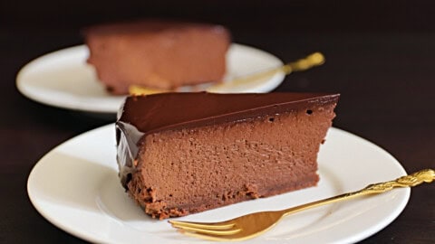 A slice of rich chocolate cheesecake with a smooth, glossy chocolate topping sits on a white plate with a gold fork beside it. Another identical slice is blurred in the background on a similar plate. The background is dark, highlighting the dessert.