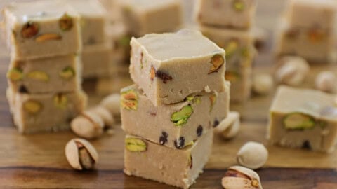 A close-up of several pieces of light brown pistachio fudge stacked on a wooden surface. The fudge is interspersed with whole pistachio nuts, and additional pistachios are scattered around the fudge on the table. The background shows more pieces of fudge.