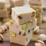 A close-up of several pieces of light brown pistachio fudge stacked on a wooden surface. The fudge is interspersed with whole pistachio nuts, and additional pistachios are scattered around the fudge on the table. The background shows more pieces of fudge.