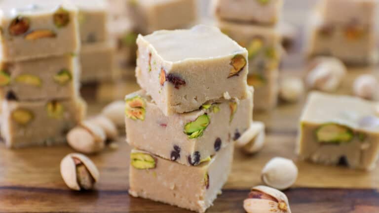 A close-up of several pieces of light brown pistachio fudge stacked on a wooden surface. The fudge is interspersed with whole pistachio nuts, and additional pistachios are scattered around the fudge on the table. The background shows more pieces of fudge.