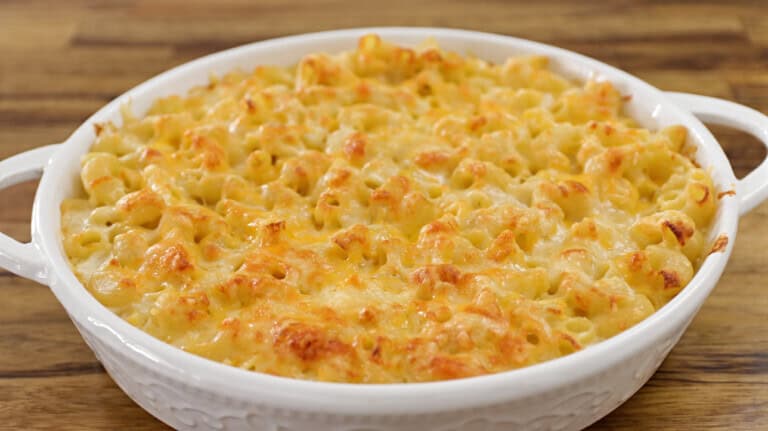 A white ceramic dish filled with golden-brown baked macaroni and cheese. The pasta is covered in melted cheese and has a slightly crispy top. The dish rests on a wooden surface, highlighting the textures and warmth of the comfort food.