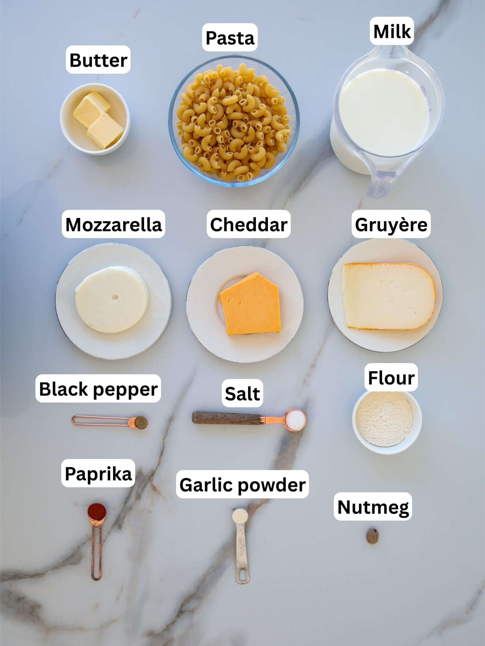 A marble surface with labeled ingredients for mac and cheese: butter, pasta, milk, mozzarella, cheddar, Gruyère, black pepper, salt, flour, paprika, garlic powder, and nutmeg, each displayed in separate dishes or spoons.