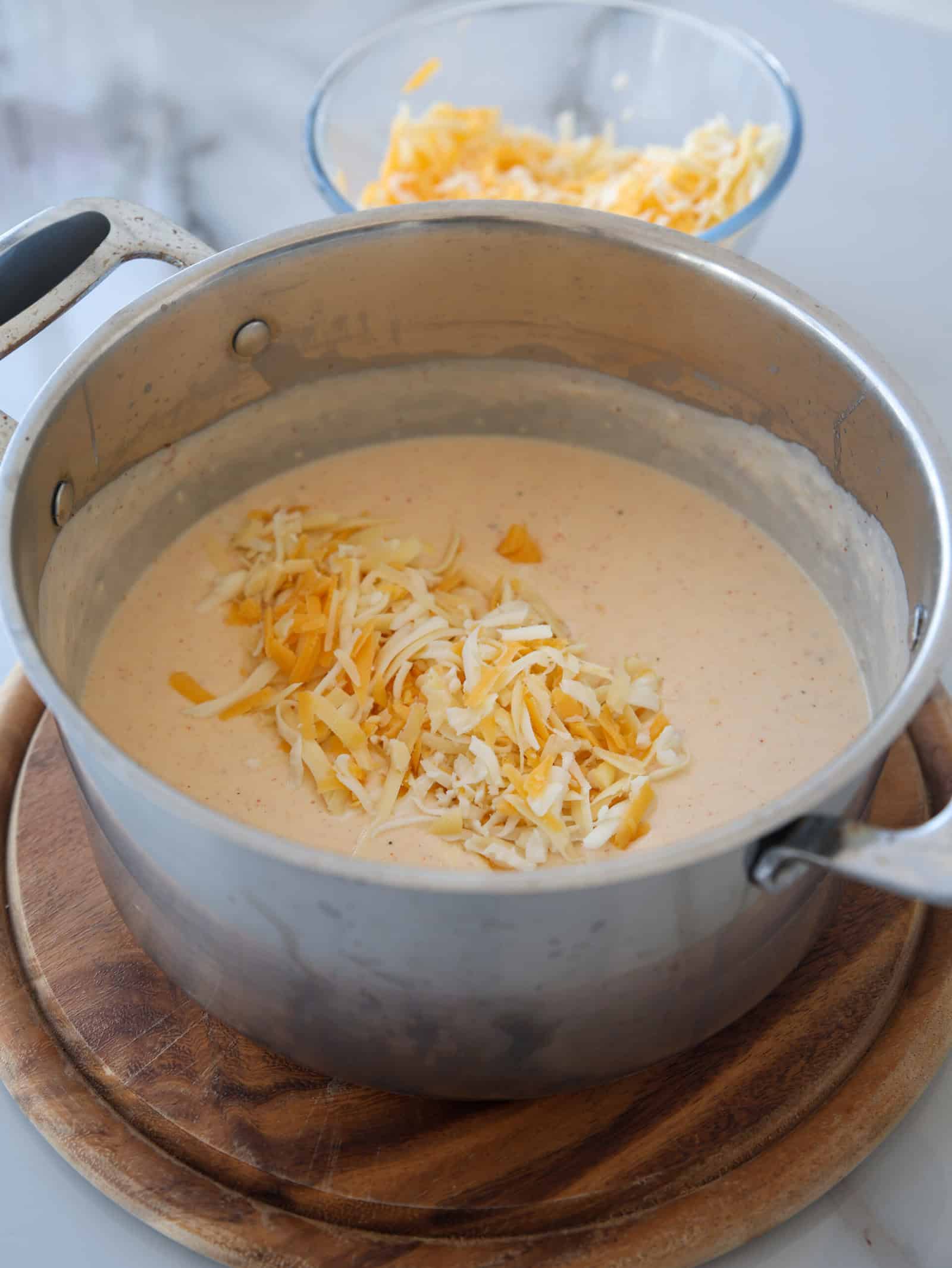 A metal pot filled with creamy cheese sauce sits on a wooden trivet. Shredded cheese is on top of the sauce, with a glass bowl of more shredded cheese in the background.