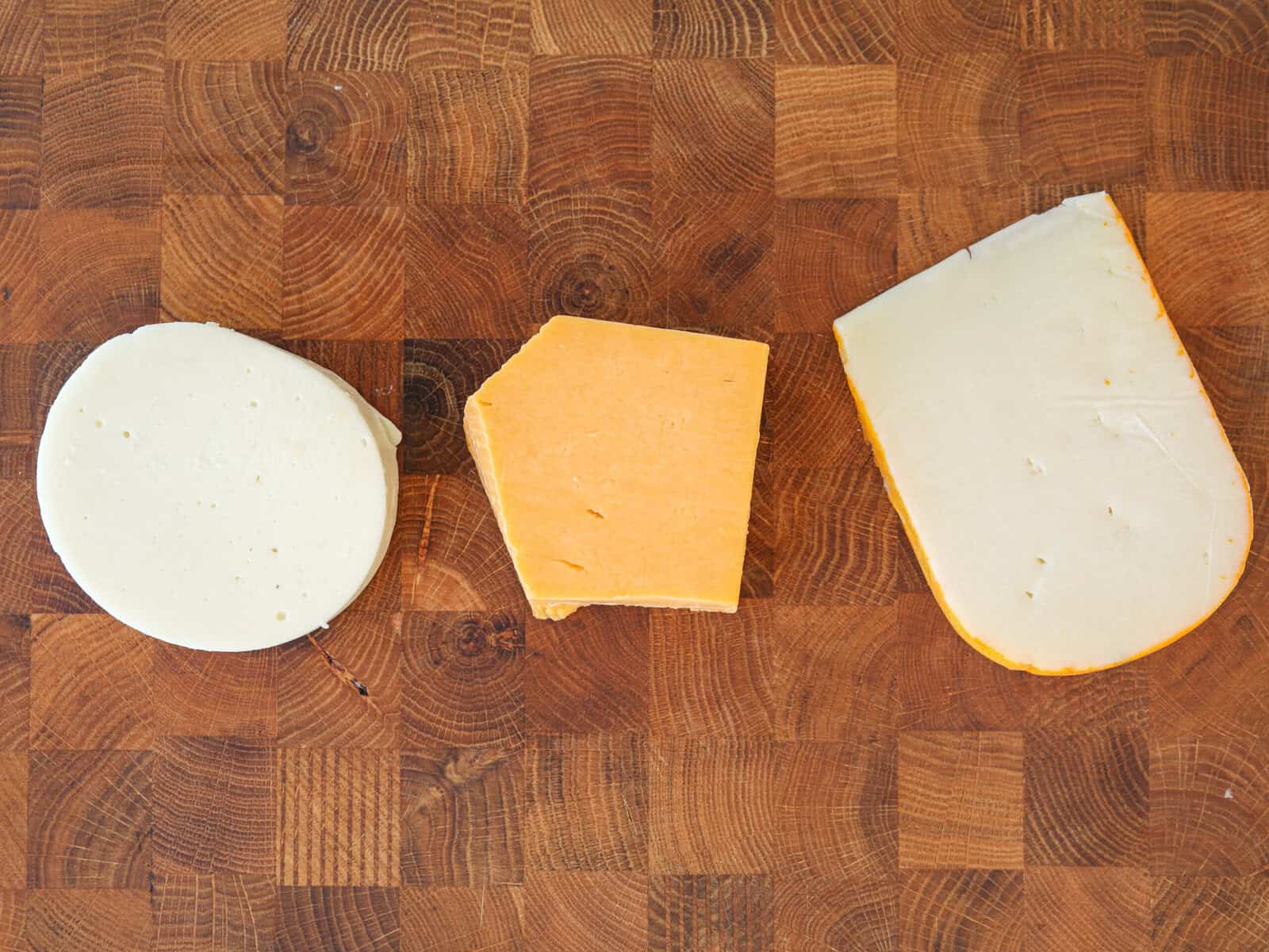 Three slices of cheese—Mozzarella (left), cheddar (center), and Gruyère  (right)—are arranged in a row on a wooden cutting board with a checkered pattern.