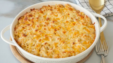 A baked dish of macaroni and cheese topped with melted, golden-brown cheese sits on a round trivet, with a fork and a checked kitchen towel nearby.