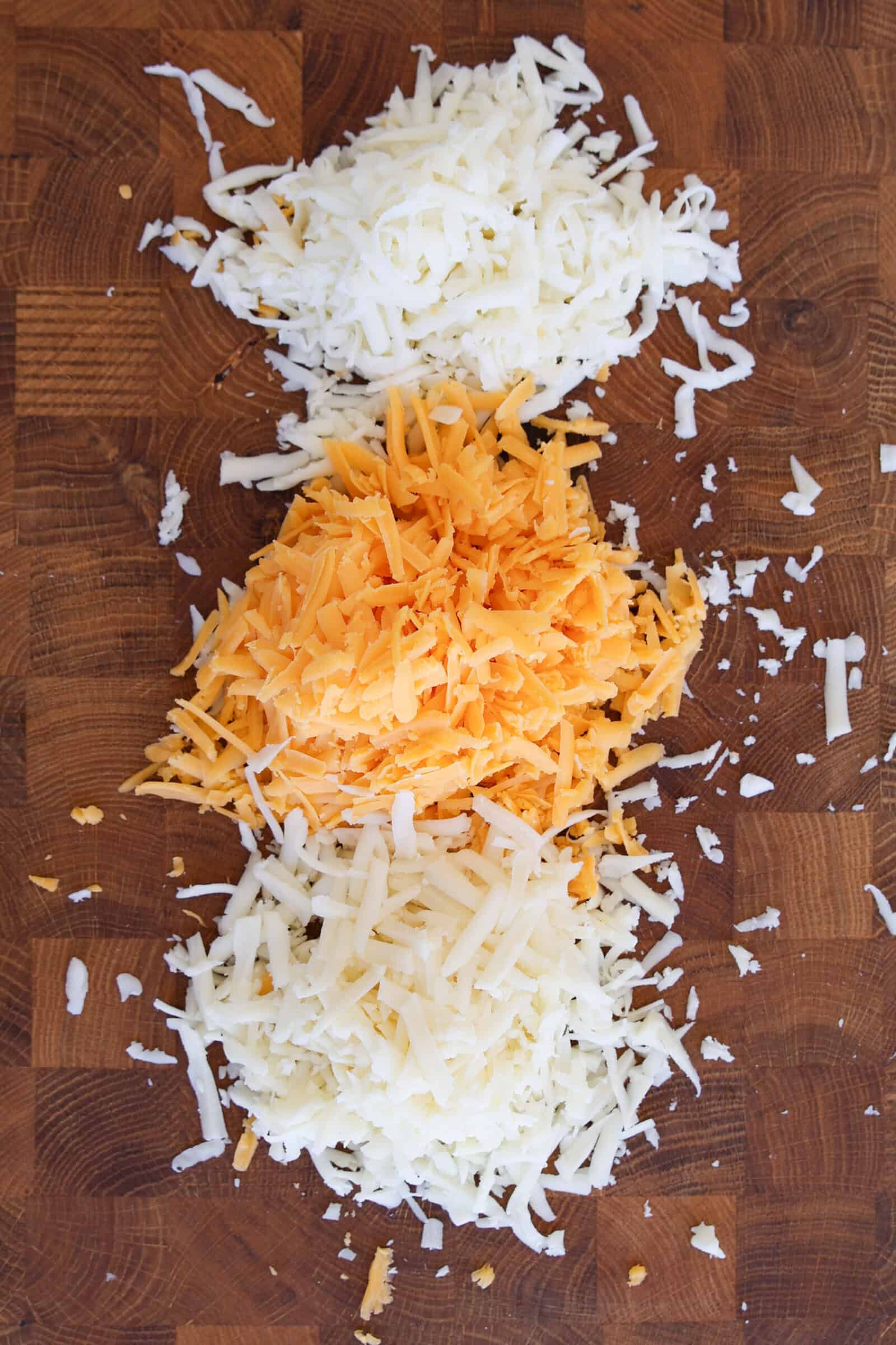 Three piles of shredded cheese—mozzarella, cheddar, and Gruyère  Jack—are arranged in a vertical line on a wooden surface.