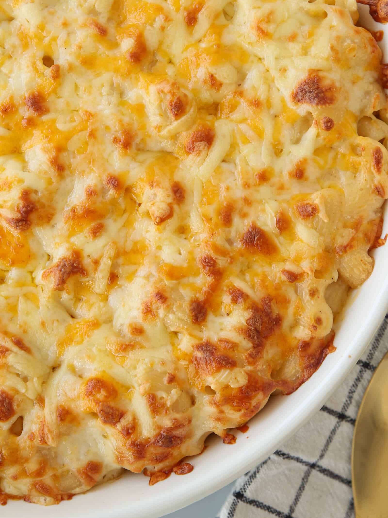 Baked macaroni and cheese in a white dish, topped with melted, golden-brown cheese. The dish rests on a checkered cloth next to a gold-colored utensil.