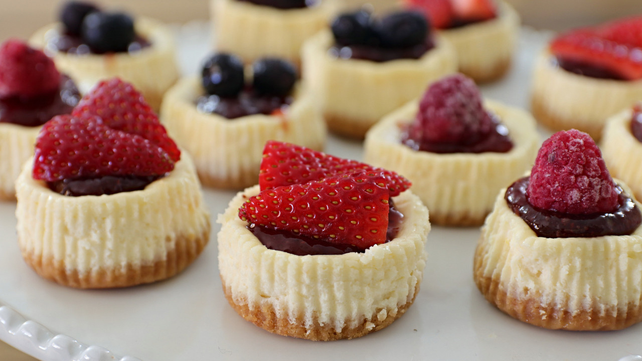 Mini cheesecakes topped with a variety of fresh fruits, including sliced strawberries, raspberries, and blueberries, are displayed on a white plate. The cheesecakes have a creamy texture with a graham cracker crust.