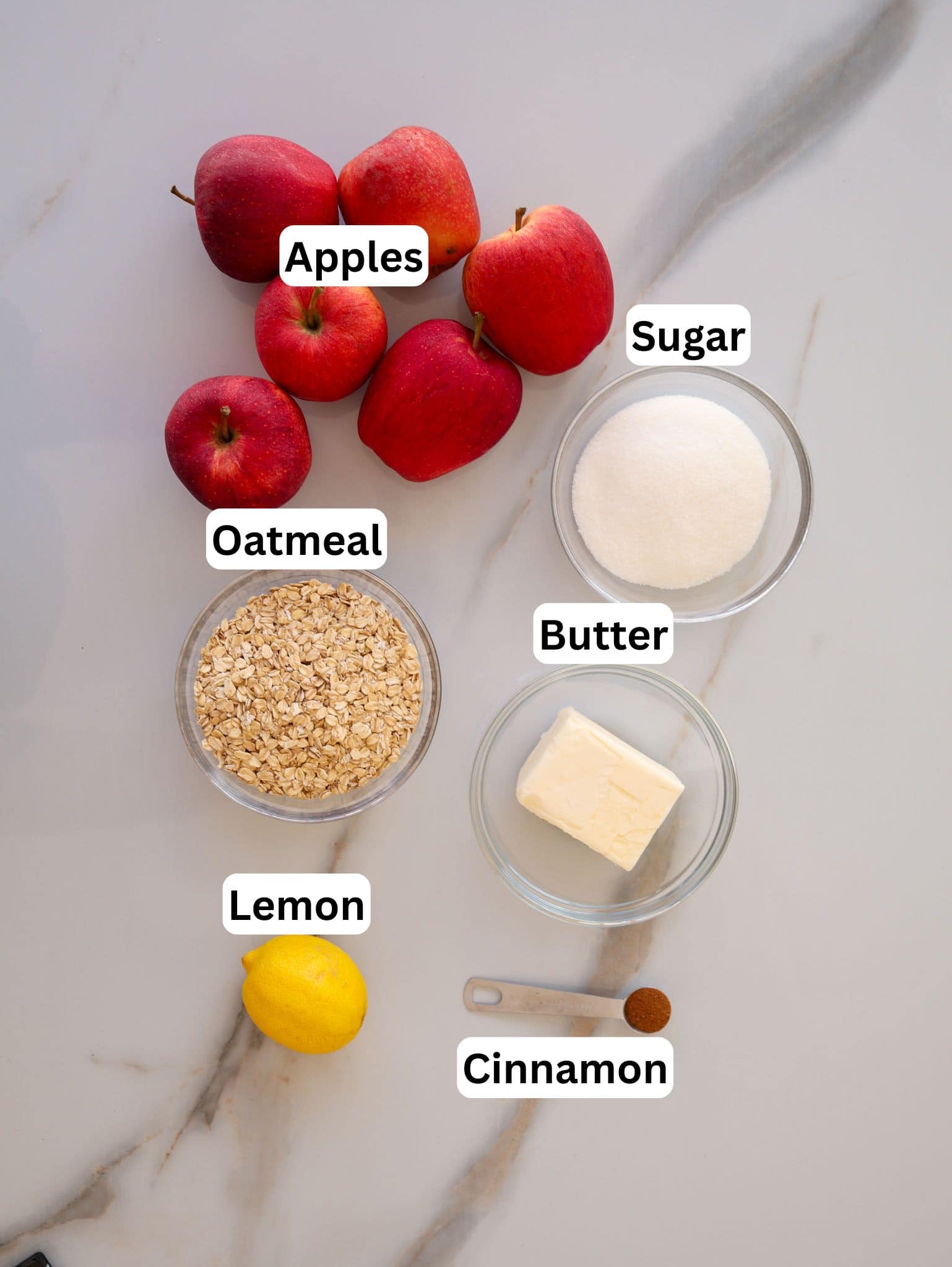 Top-down view of apples, a bowl of sugar, a bowl of oatmeal, a bowl of butter, a lemon, and a teaspoon of cinnamon on a marble surface, each ingredient labeled with white text.