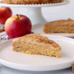 A slice of layered apple cake on a white plate, surrounded by red apples, cinnamon sticks, and a fork, with the rest of the cake and more apples in the background.